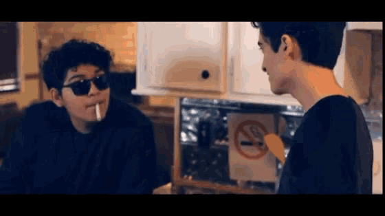 a man is smoking a cigarette next to another man who is wearing sunglasses .