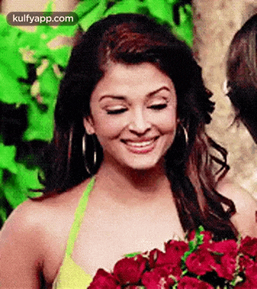 a woman in a yellow top is smiling while holding a bouquet of red roses .
