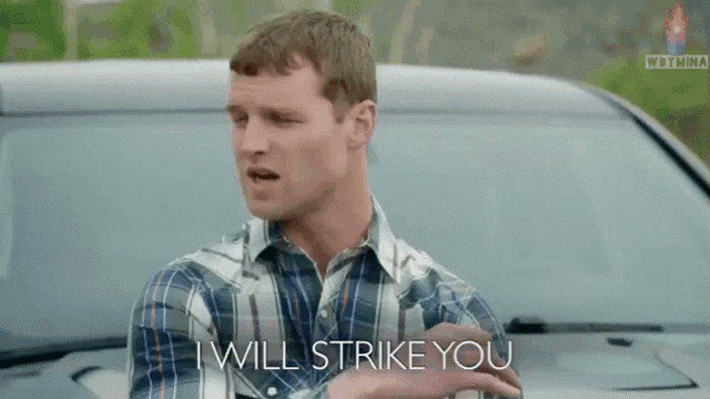 a man in a plaid shirt stands in front of a car and says i will strike you