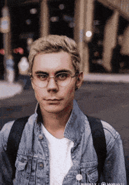 a young man wearing glasses and a denim jacket stands in front of a building