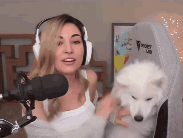 a woman wearing headphones is holding a white dog in front of a microphone while sitting in a secret lab chair