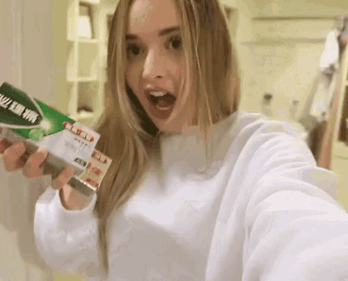 a woman in a white sweatshirt is holding a box of toothpaste in her hands