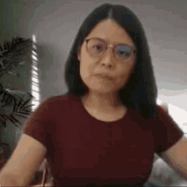 a woman wearing glasses and a maroon t-shirt is sitting in front of a plant .