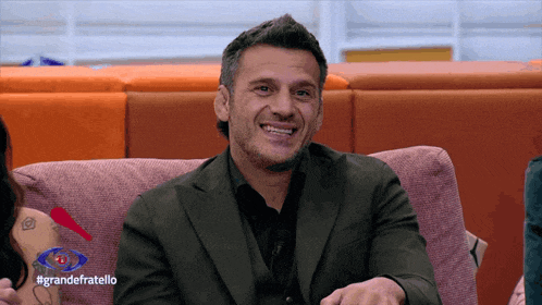 a man in a suit sits on a couch with the hashtag grandefratello on the bottom right