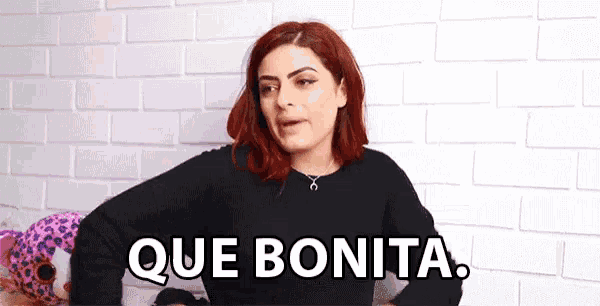 a woman with red hair is saying que bonita in front of a white brick wall
