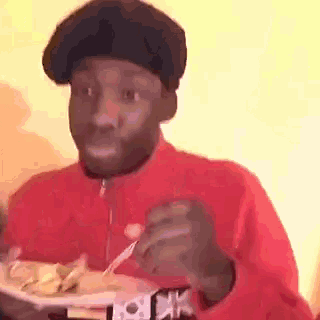 a man in a red jacket and black hat is eating a plate of food with a fork .