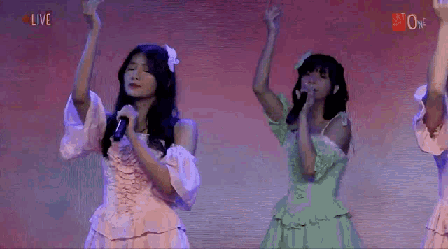 two girls are singing into microphones on a stage with the word live in the corner .