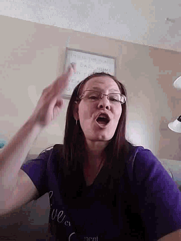 a woman wearing glasses and a purple shirt has her mouth open and is pointing at something