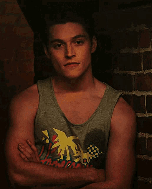 a man wearing a tank top with a palm tree on it is standing with his arms crossed in front of a brick wall