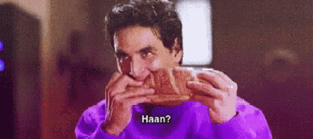 a man in a purple sweater is eating a piece of bread and says `` haan '' .