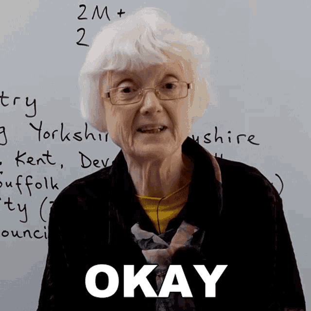 an elderly woman stands in front of a white board that says " okay "