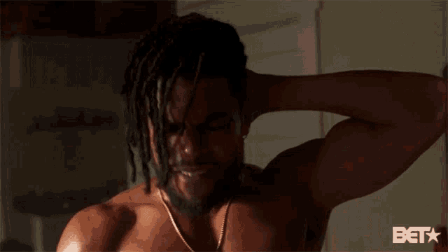 a shirtless man with dreadlocks and a necklace holds his head in pain