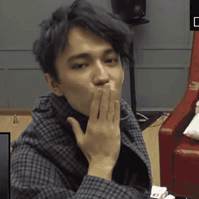 a young man covering his mouth with his hand while wearing a plaid jacket
