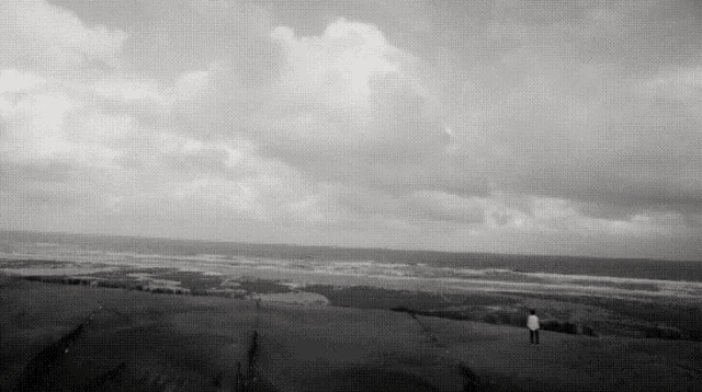 a black and white photo of a man standing on top of a hill overlooking the ocean