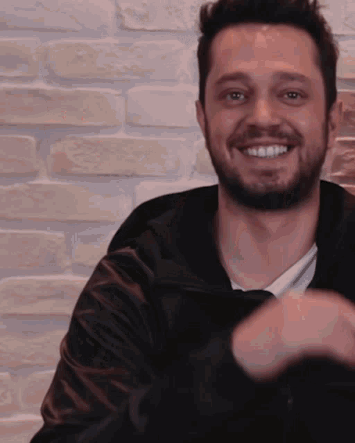 a man wearing a black jacket is smiling in front of a white brick wall