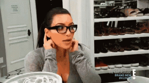 a woman wearing glasses and a gray shirt is sitting in front of a closet filled with shoes .