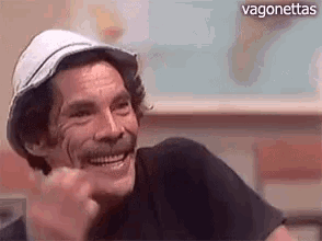 a man wearing a hat and a black shirt is smiling and pointing at the camera .