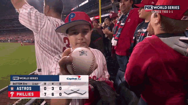 a young boy holds a baseball in front of a scoreboard that says fox world series series tied 1-1