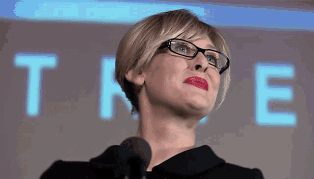 a woman wearing glasses and red lipstick stands in front of a microphone in front of the word time