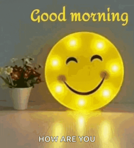 a smiley face is sitting on a table next to a vase of flowers and says `` good morning how are you '' .