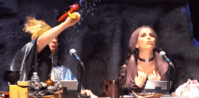a woman with purple hair is sitting at a table with two microphones and a bottle of water