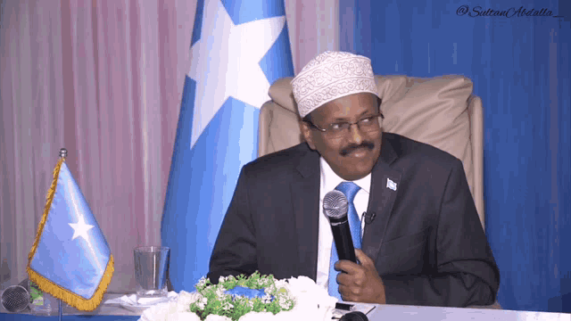 a man sitting at a table with a microphone in front of a flag