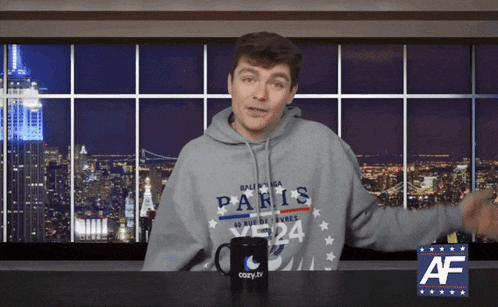 a man wearing a paris sweatshirt stands in front of a cozy.tv mug