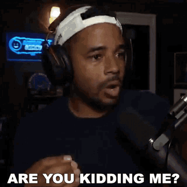 a man wearing headphones and a headband is talking into a microphone and asking are you kidding me .