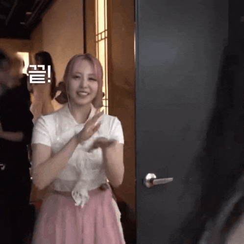 a woman in a pink skirt is standing in front of a door and clapping her hands .