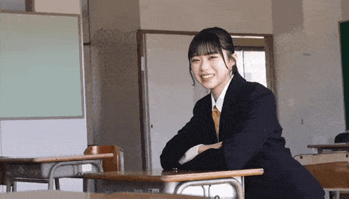 a girl in a suit sits at a desk in a classroom smiling