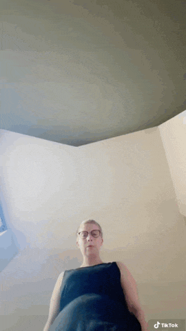 a woman wearing glasses and a black tank top is standing in a room with a tiktok logo on the bottom right