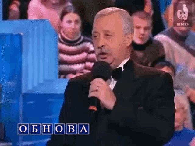 a man in a tuxedo is holding a microphone in front of a crowd and the word obnova is on the screen