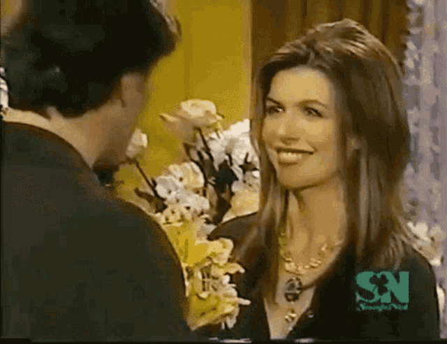 a woman is smiling at a man in front of flowers and the letters sn are on the bottom of the screen