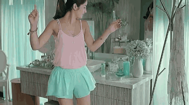 a woman in a pink tank top and blue shorts is dancing in a bathroom in front of a mirror .