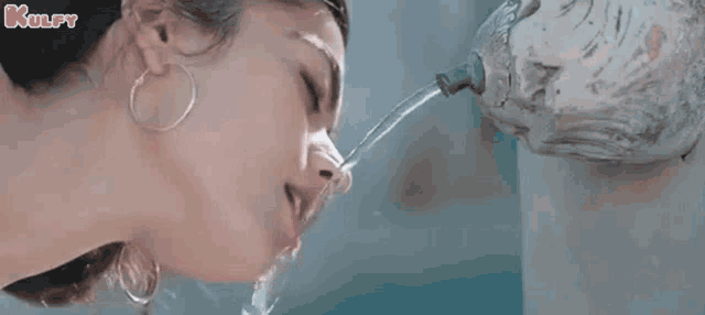 a woman drinking water from a fountain with kulfy written on the bottom right