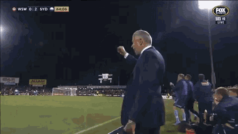 a man in a suit stands on a soccer field in front of a fox sports logo
