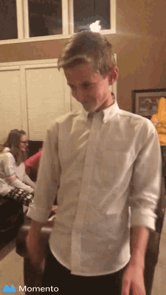 a boy in a white shirt is standing in a living room with a momento icon on the bottom right
