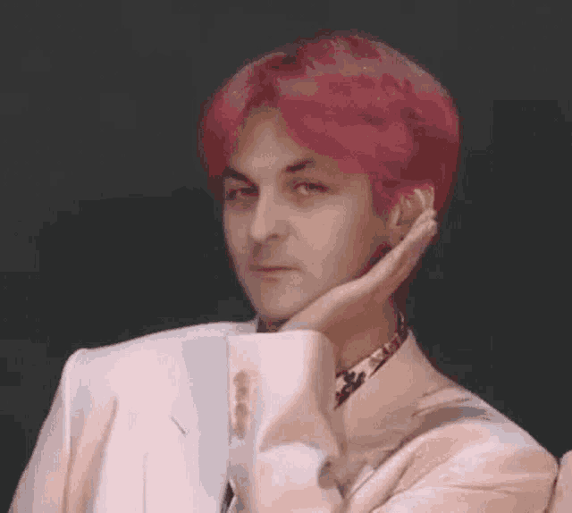 a man with pink hair is wearing a white suit and touching his face with his hand .