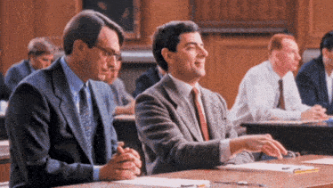 a man in a suit and tie sits next to another man at a desk