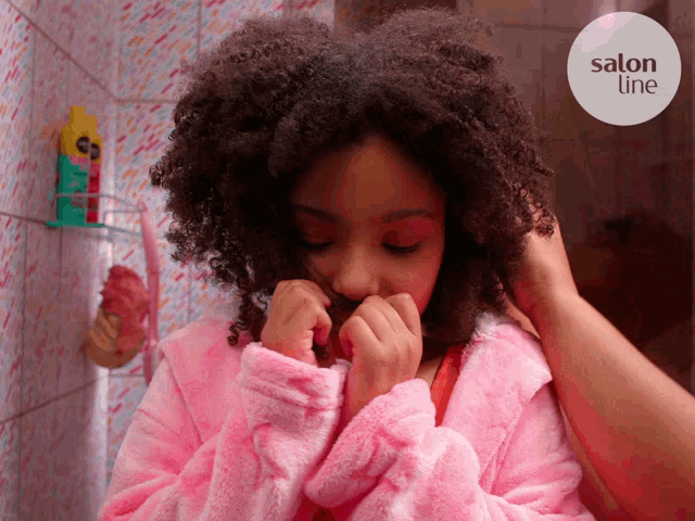 a girl in a pink robe is looking at herself in a mirror with a salon title sticker behind her