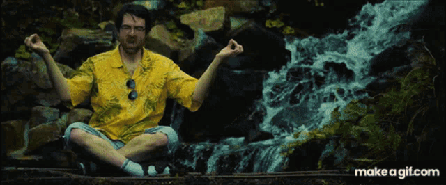 a man in a yellow shirt sits in front of a waterfall with his arms outstretched