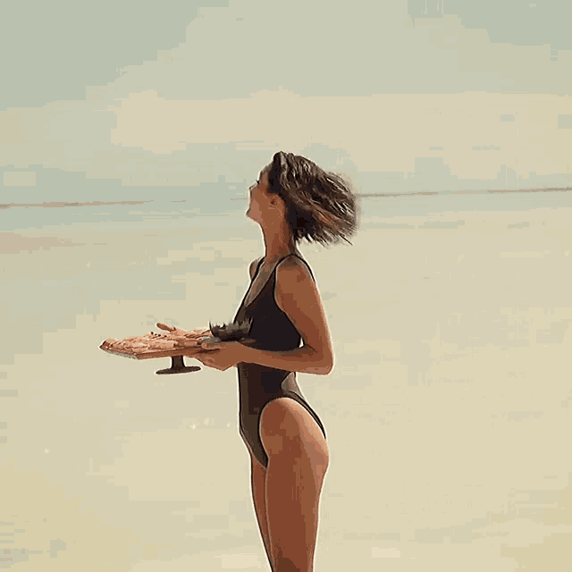 a woman in a black swimsuit holds a tray of food on the beach