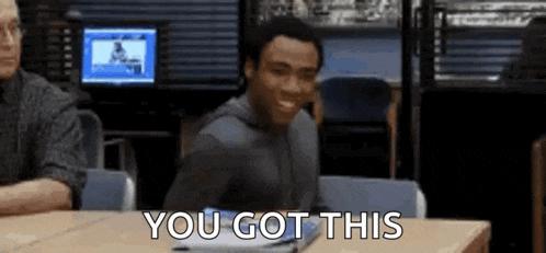 a man is sitting at a table with a laptop and smiling while someone says `` you got this '' .