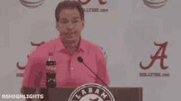 a man in a pink shirt stands at a podium with a coca cola bottle in his hand