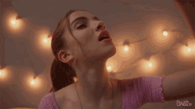 a woman in a pink top is dancing in front of a string of lights and the words brattv are on the bottom right