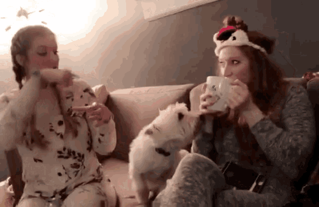 two girls are sitting on a couch drinking from cups and a dog is looking on
