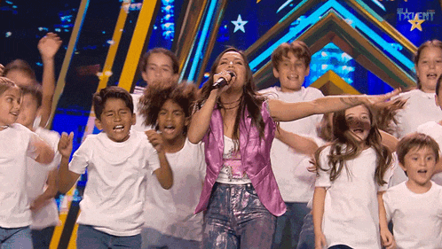 a woman in a pink jacket sings into a microphone with a group of children behind her