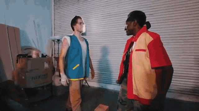 a man in a blue vest is standing next to a man in a red shirt .