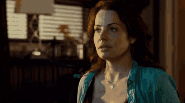 a woman in a blue shirt and necklace is standing in a dark room looking at something .