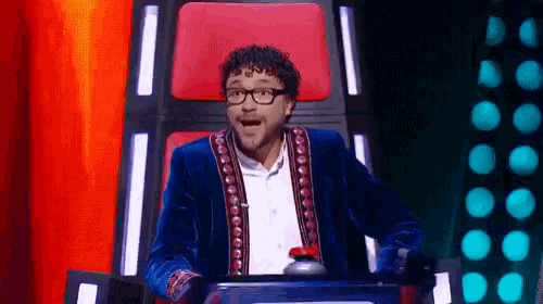 a man wearing glasses and a blue jacket is sitting in a chair with a red seat behind him .